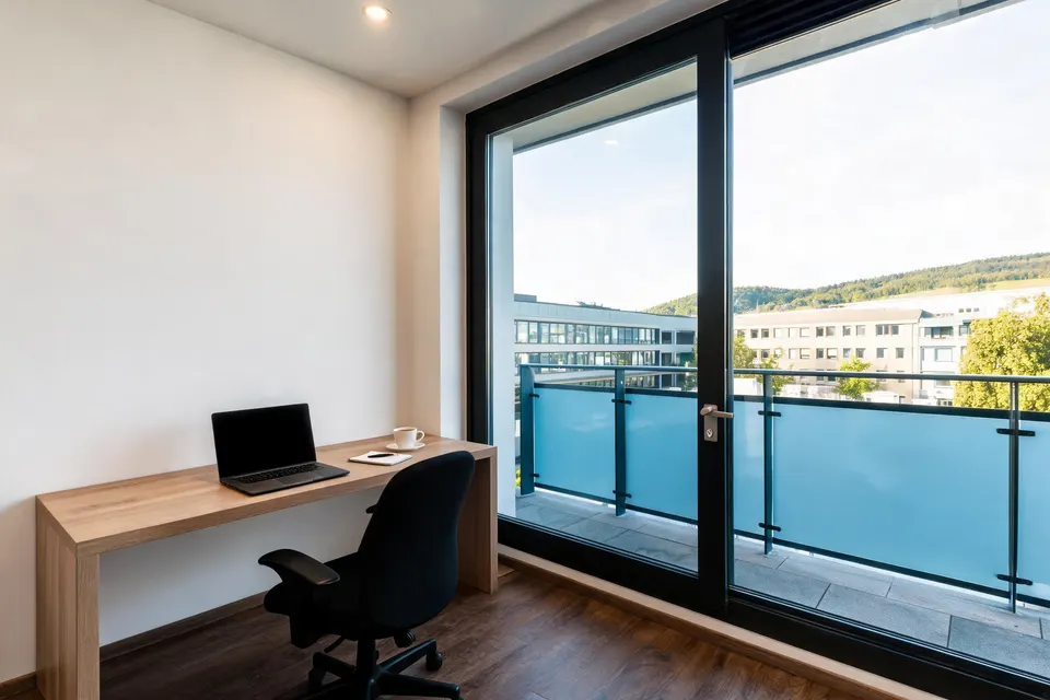 Schreibtisch mit Laptop und Kaffee an einer schwarz gerahmten Balkontür mit blauem Glasgeländer, Blick auf Flachbauten und Hügel (Symbolbild im Stil des Hauses)
