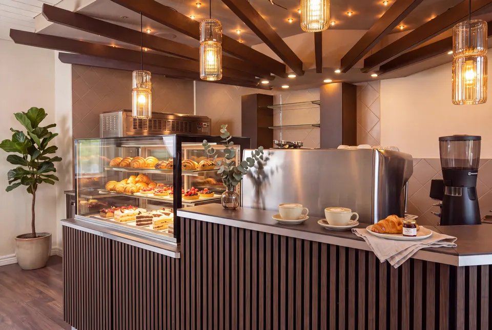 Theke des CaféDeRia mit Vitrine voller Gipfeli, Tartes und Torten, zwei Cappuccini und ein Croissant auf der Theke, Eukalyptus in der Vase, Espressomaschine und Fächer aus dunklen Balken (Symbolbild auf Basis einer Aufnahme des Hauses)