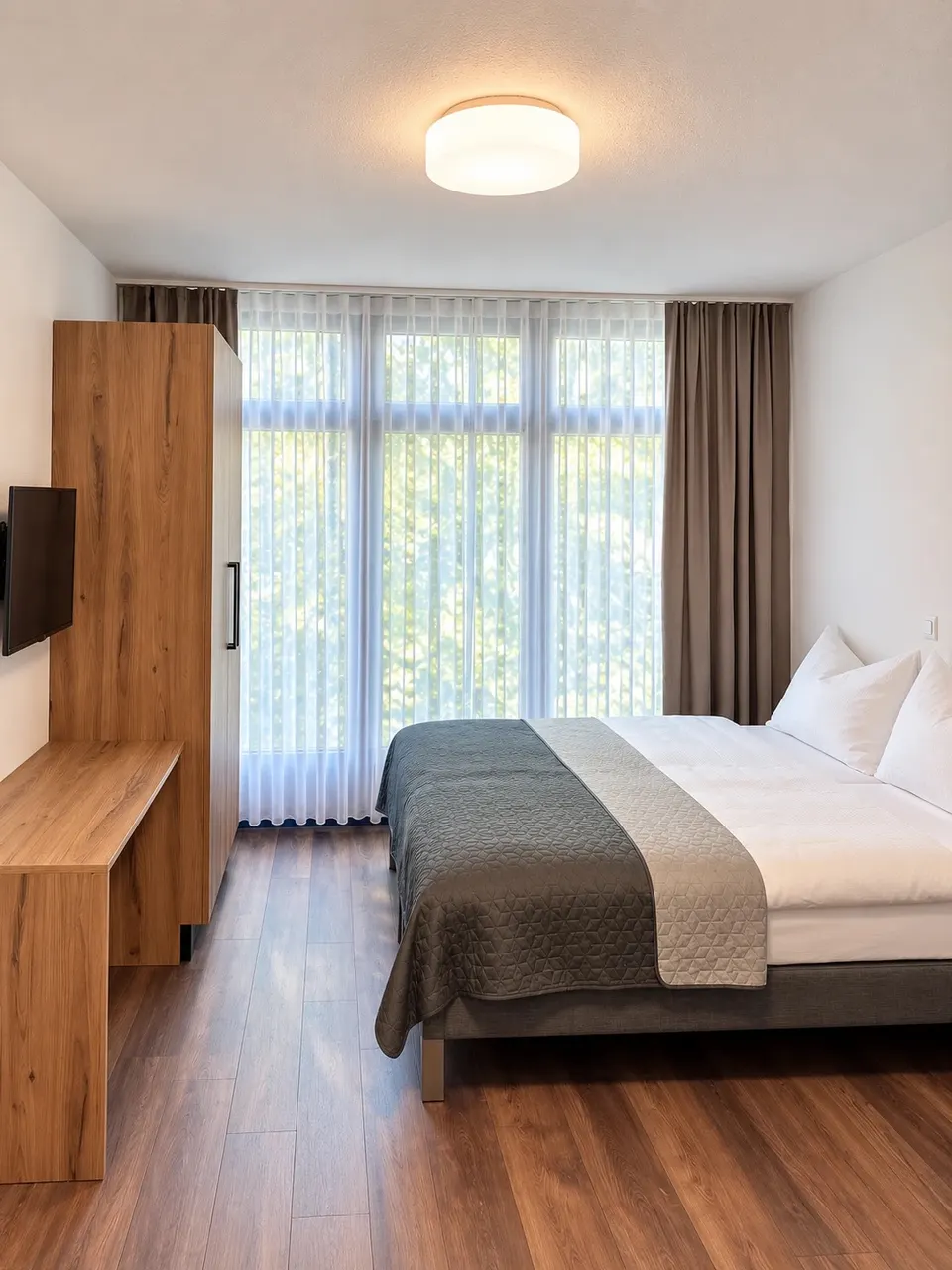 Studio ohne Balkon: Doppelbett vor einer breiten Fensterfront mit Vorhängen, Schrank, Schreibtisch und Wand-TV
